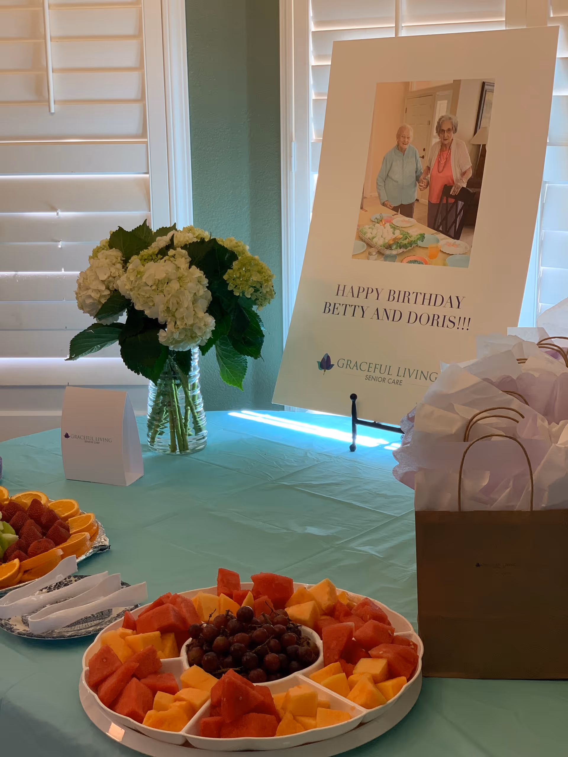 A table set up for a birthday celebration with a platter of assorted fresh fruit including watermelon, cantaloupe, and grapes, a vase of white hydrangeas, gift bags with tissue paper, and a large sign that reads 'Happy Birthday Betty and Doris!!!' with a photo of two elderly women holding hands. The sign also displays the Graceful Living Senior Care logo.