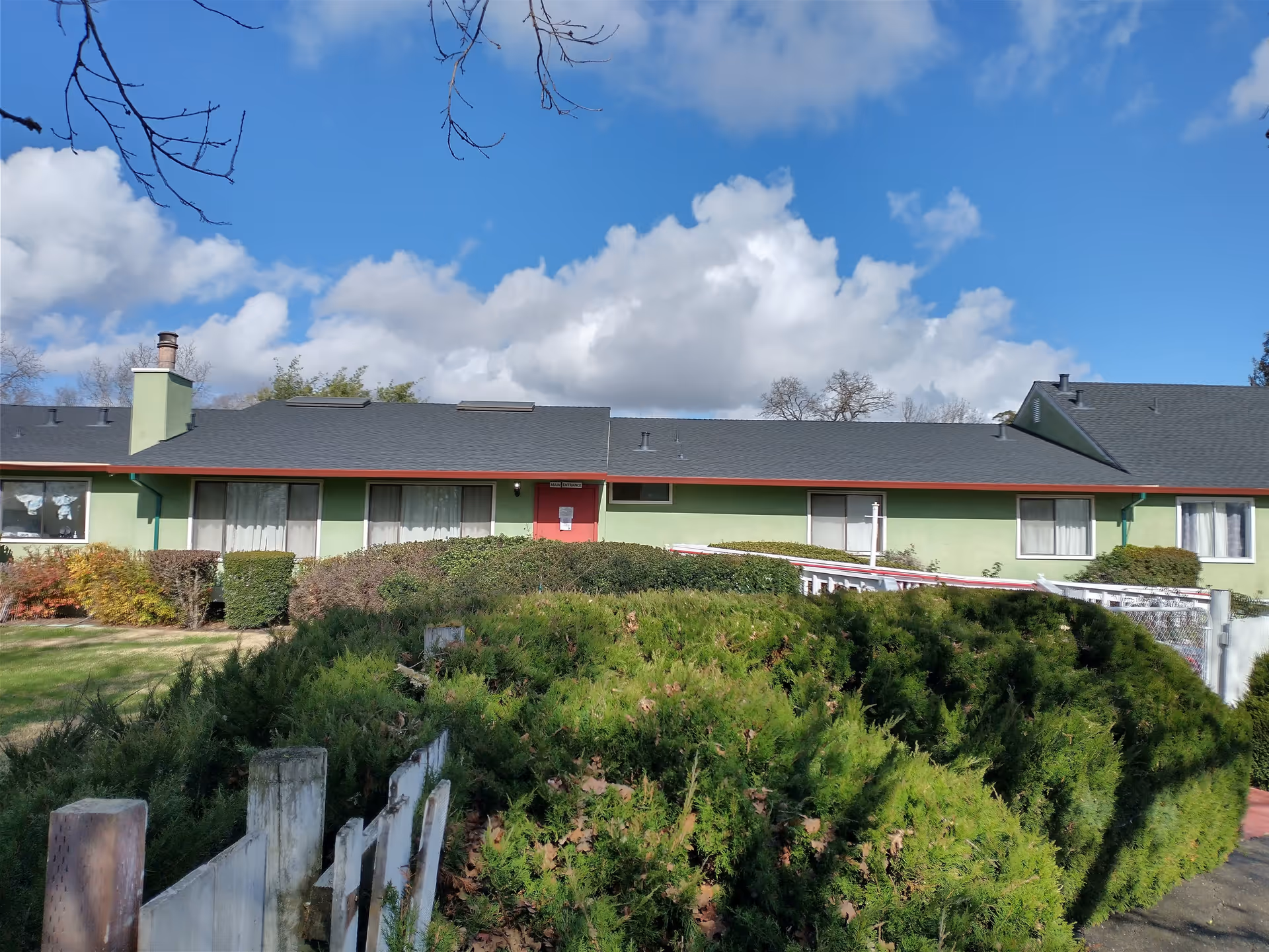 Exterior view of a single-story building with green walls and a dark roof under a partly cloudy blue sky. The building is surrounded by bushes and a small white fence in the foreground.