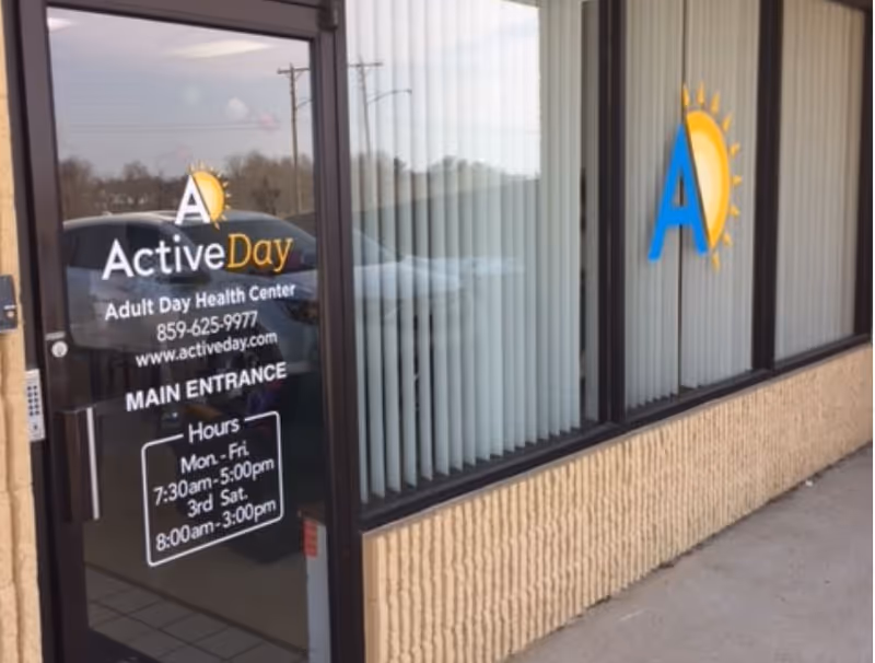 Glass door entrance to Active Day Adult Day Health Center with business hours and contact information displayed on the door. Adjacent windows have vertical blinds and a logo with a blue letter A and a yellow sun.