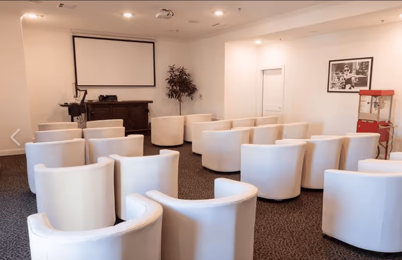 A small theater or presentation room with several rows of white cushioned chairs facing a projector screen. There is a wooden cabinet with electronic equipment beneath the screen, a potted plant in the corner, a popcorn machine on the right side, and a black and white framed photograph on the wall.