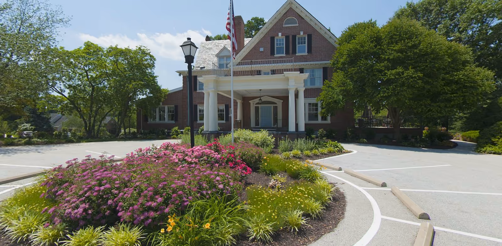 Front exterior view of a large brick building with white columns at the entrance, surrounded by a landscaped garden with colorful flowers and green trees under a blue sky.