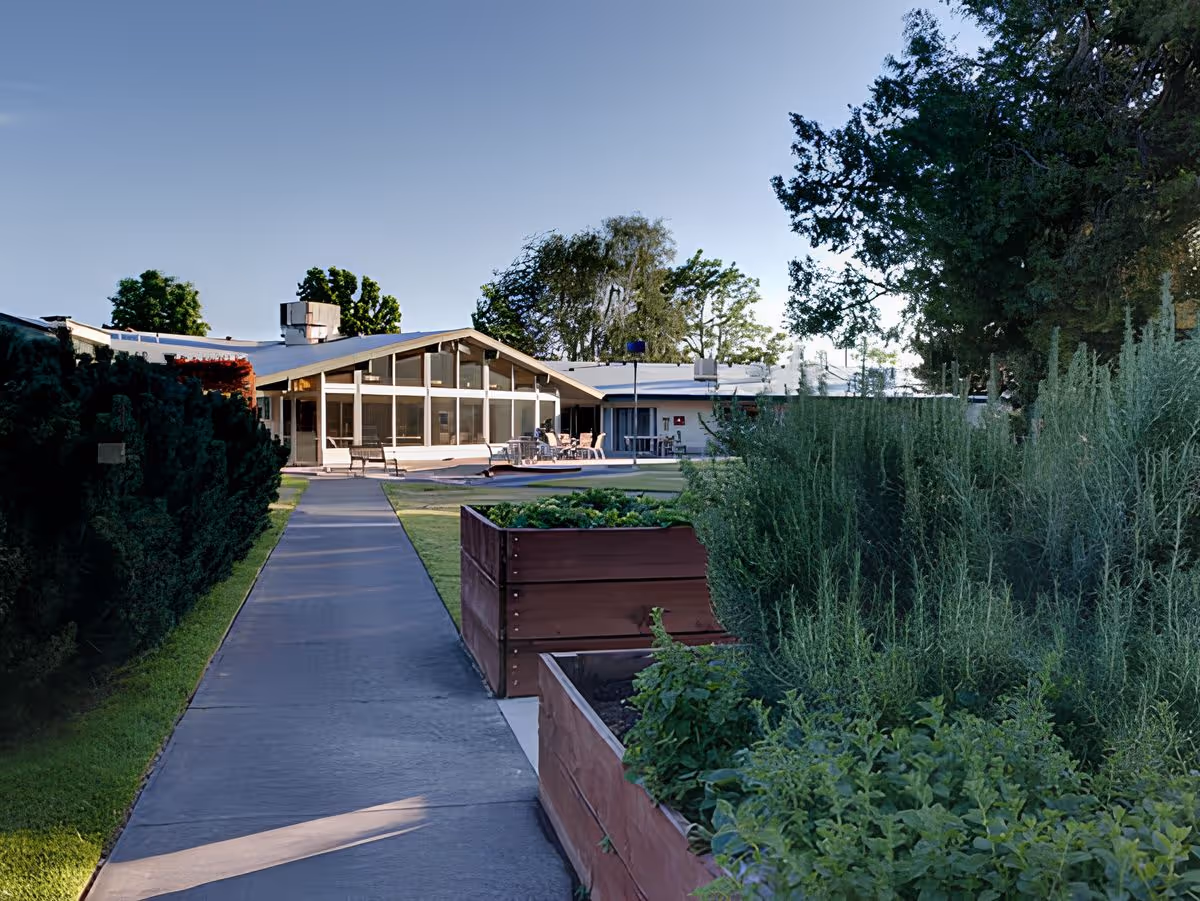 A paved walkway lined with raised garden beds and greenery leading to the entrance of a single-story senior living facility building.