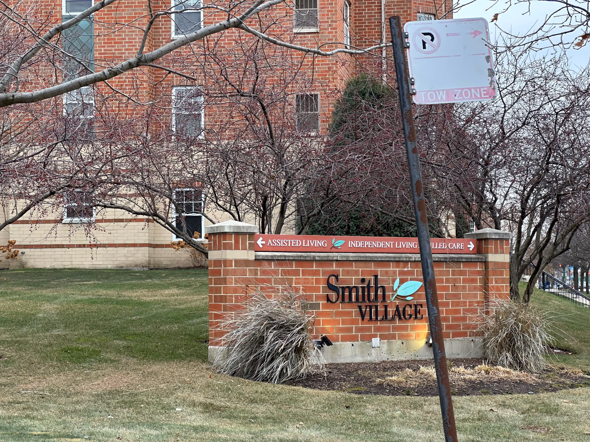Brick sign for Smith Village senior living facility with directional arrows for Assisted Living, Independent Living, and Skilled Care. The sign is surrounded by grass, bushes, and leafless trees, with a brick building in the background and a rusty no parking sign in the foreground.