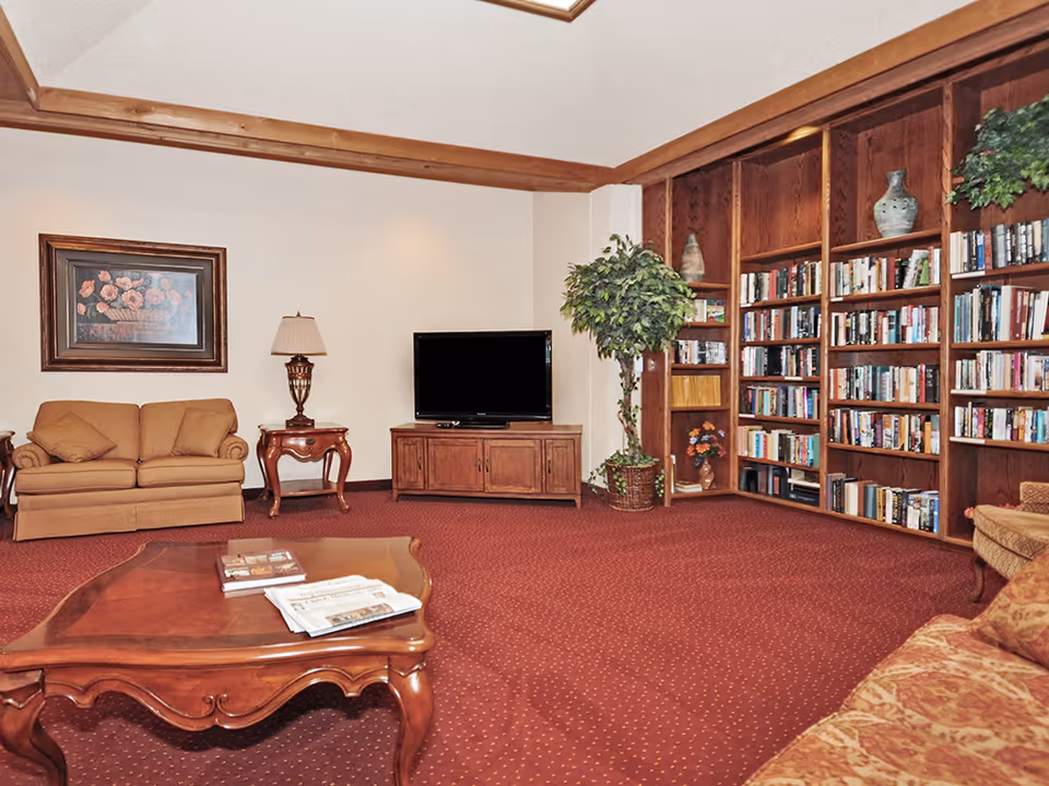 A cozy living room with a red carpet, wooden coffee table with books and newspapers, beige sofas, a wooden TV stand with a flat-screen TV, a side table with a lamp, a large potted plant, and a wooden bookshelf filled with books and decorative items.