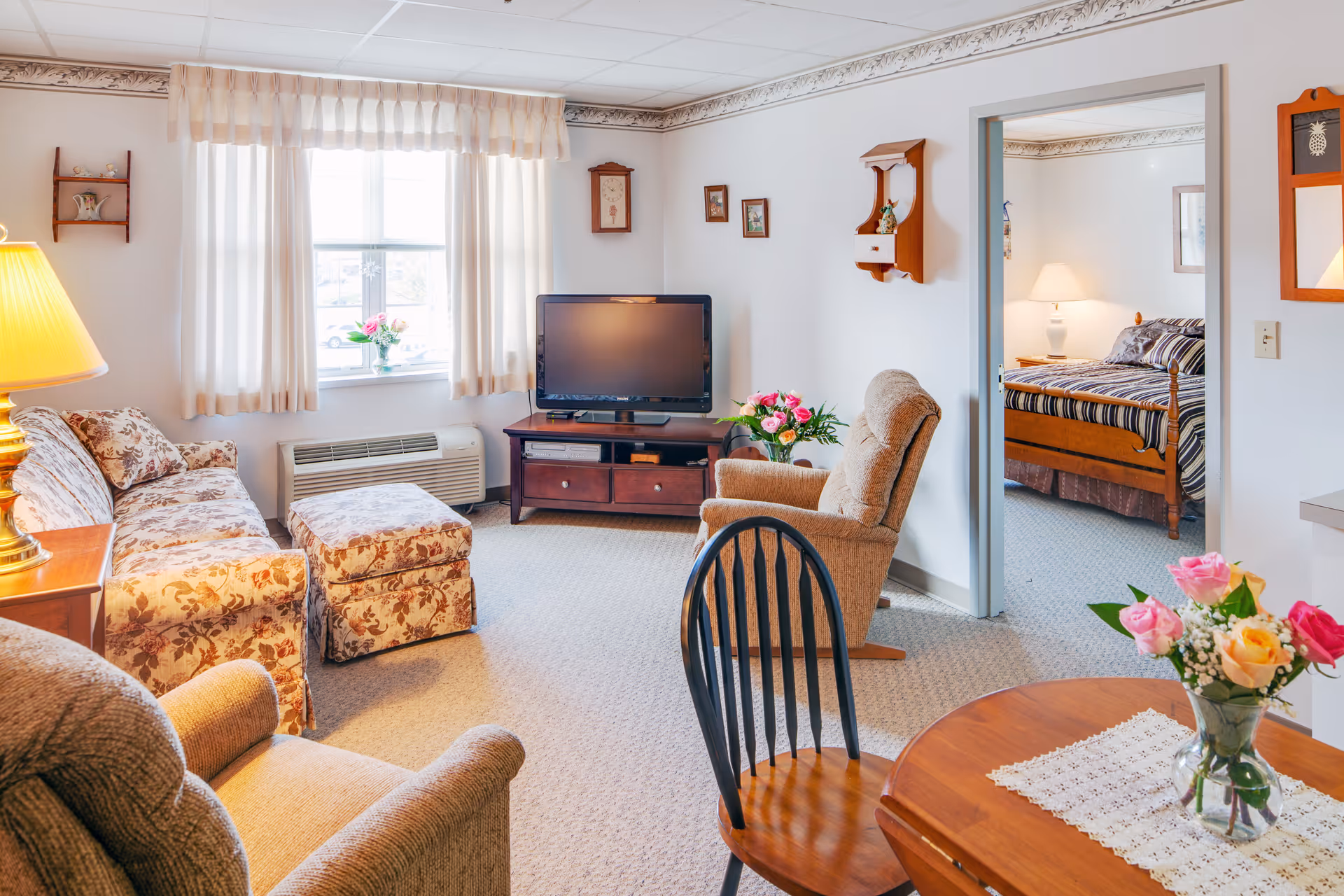 A cozy living room with a floral patterned sofa and matching ottoman, a beige recliner, a wooden TV stand with a flat screen TV, and a window with sheer curtains letting in natural light. A round wooden table with a vase of colorful flowers is in the foreground. Through an open doorway, a bedroom with a wooden bed frame and striped bedding is visible.