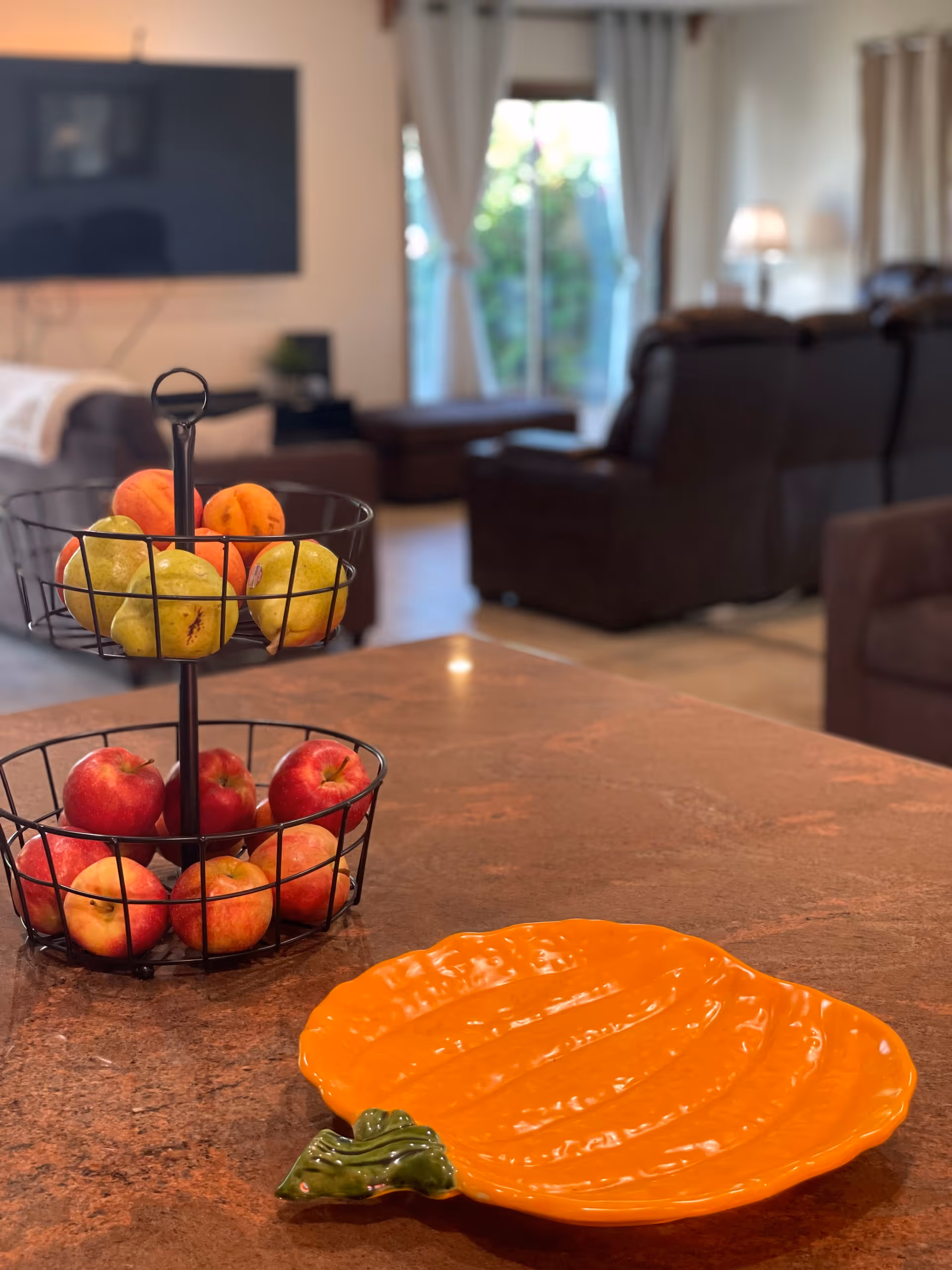 A close-up view of a kitchen countertop with a two-tiered wire fruit basket holding apples and pears, and an orange pumpkin-shaped plate. In the background, there is a living room area with brown leather sofas, a large flat-screen TV mounted on the wall, and a sliding glass door with curtains letting in natural light.