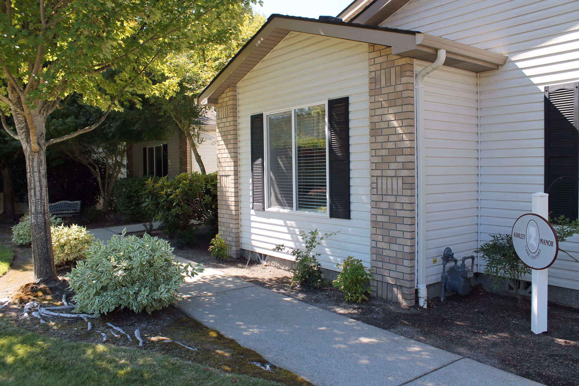 Exterior view of a single-story building with white siding and brick accents, black window shutters, and a sidewalk leading past landscaped bushes and trees. A sign near the building reads 'Ashley Manor'.