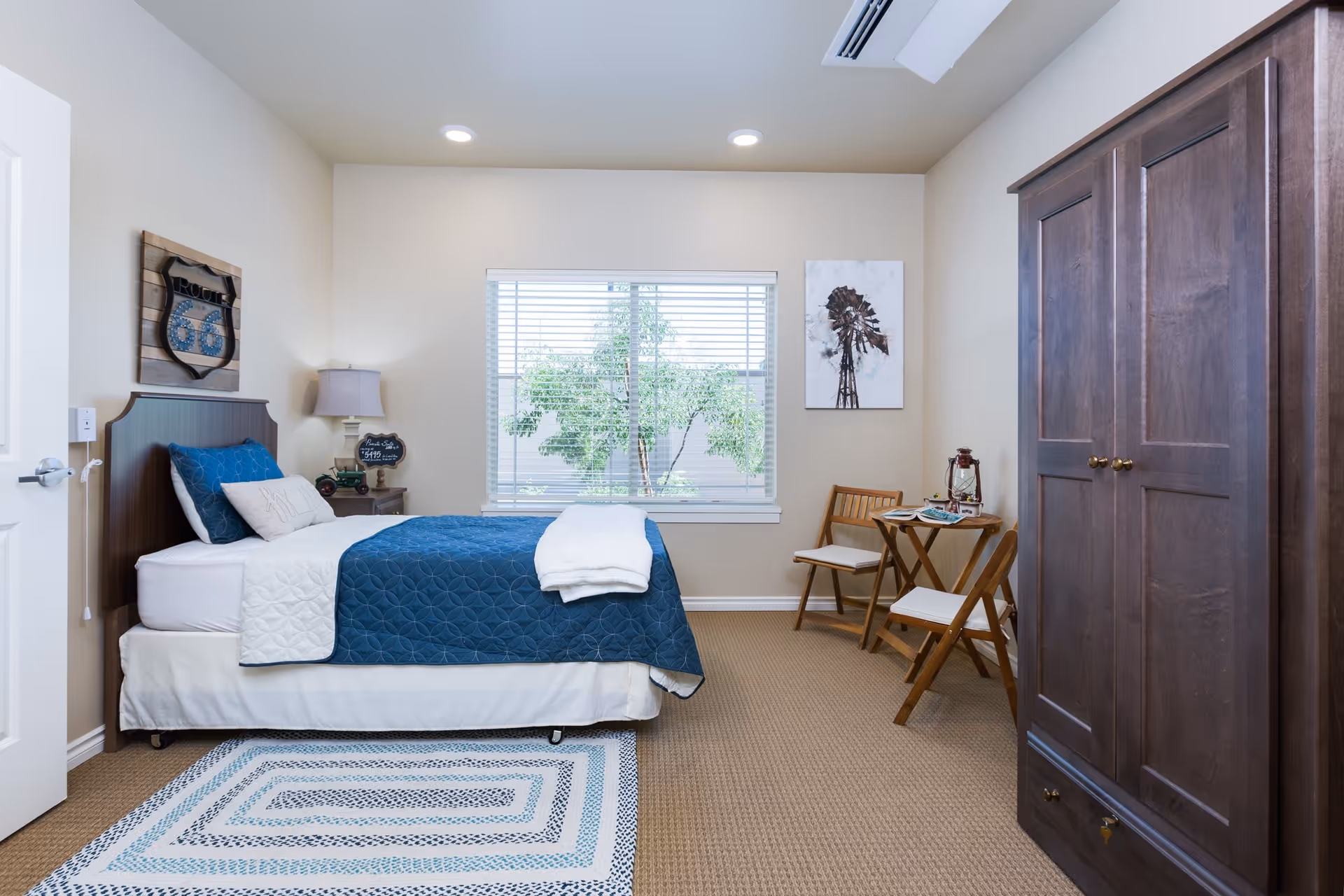 A neatly arranged bedroom with a single bed made with white and blue bedding, a wooden headboard, and a small bedside table with a lamp and decorative items. There is a window with blinds letting in natural light, a framed picture of a windmill on the wall, two wooden chairs with a small table between them, and a large wooden wardrobe. The room has beige walls and carpeted floor with a blue and white patterned rug.