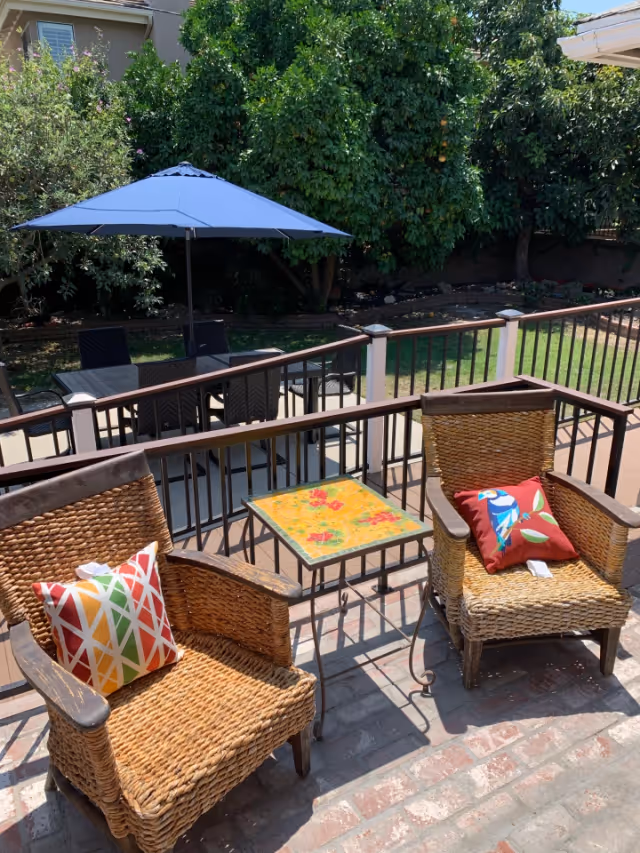 Sunlit patio with two wicker chairs topped with colorful pillows and a small floral side table, overlooking a fenced yard with a dining table and blue umbrella.