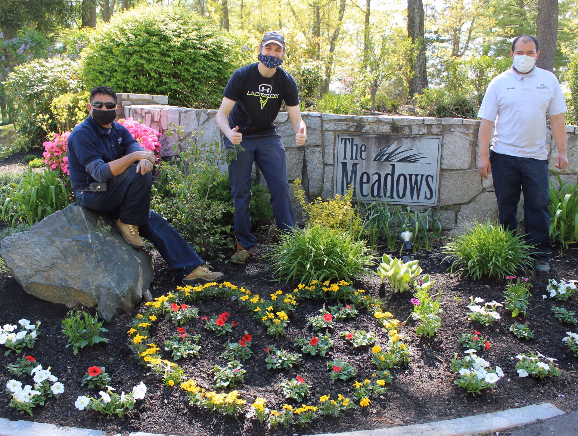 Three men wearing face masks posing outdoors in front of a stone sign that reads 'The Meadows'. They are surrounded by a garden with colorful flowers arranged in a heart shape and the word 'Thank You' spelled out in flowers.