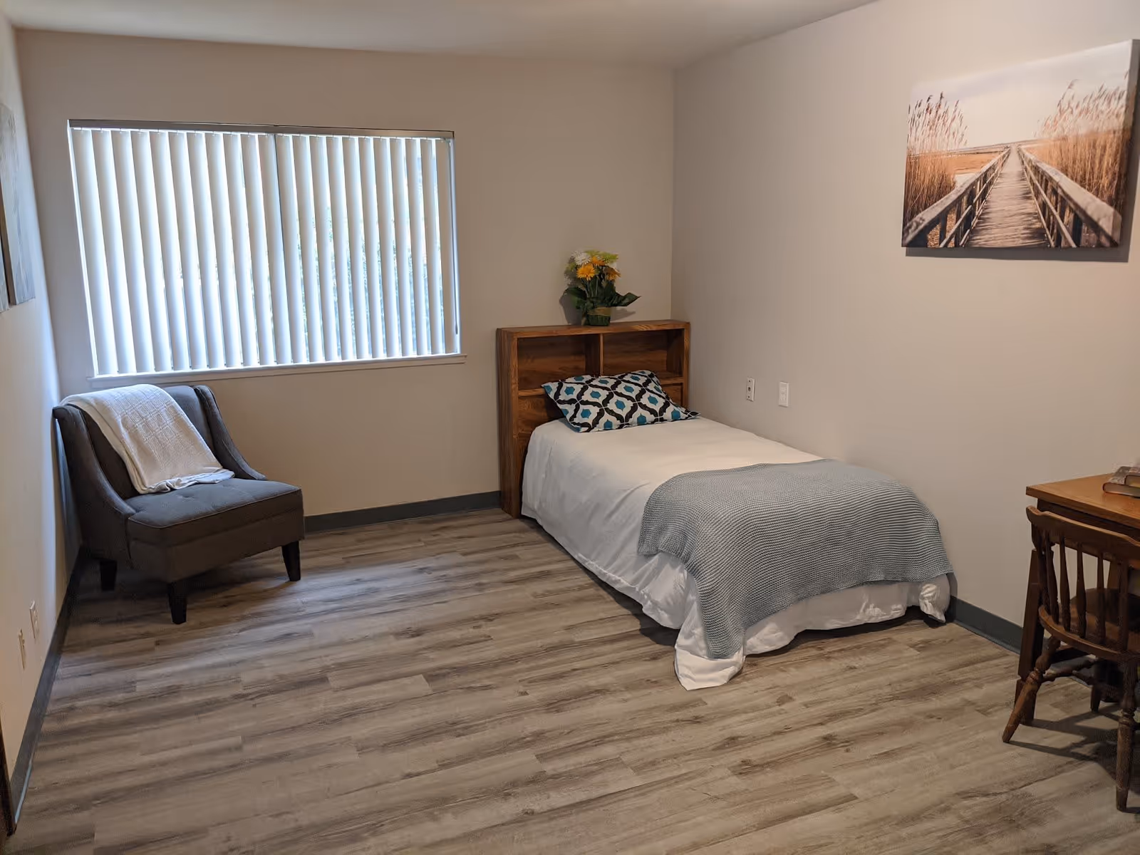 Simple bedroom with a single bed, armchair, desk, window with vertical blinds, and wall artwork.