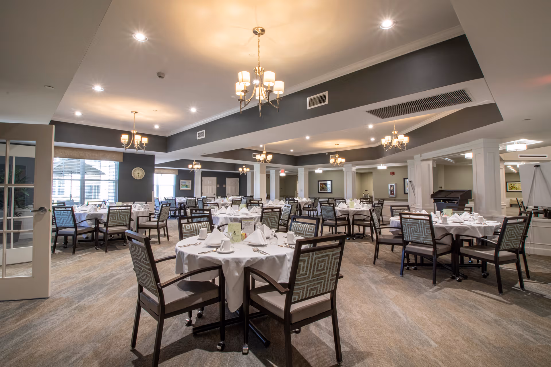 A spacious dining room with multiple round tables covered with white tablecloths, each set with white napkins, cups, and silverware. The room features modern chandeliers hanging from a ceiling with recessed lighting, large windows allowing natural light, and patterned chairs around the tables. The walls are painted in neutral tones with some artwork and a clock visible.