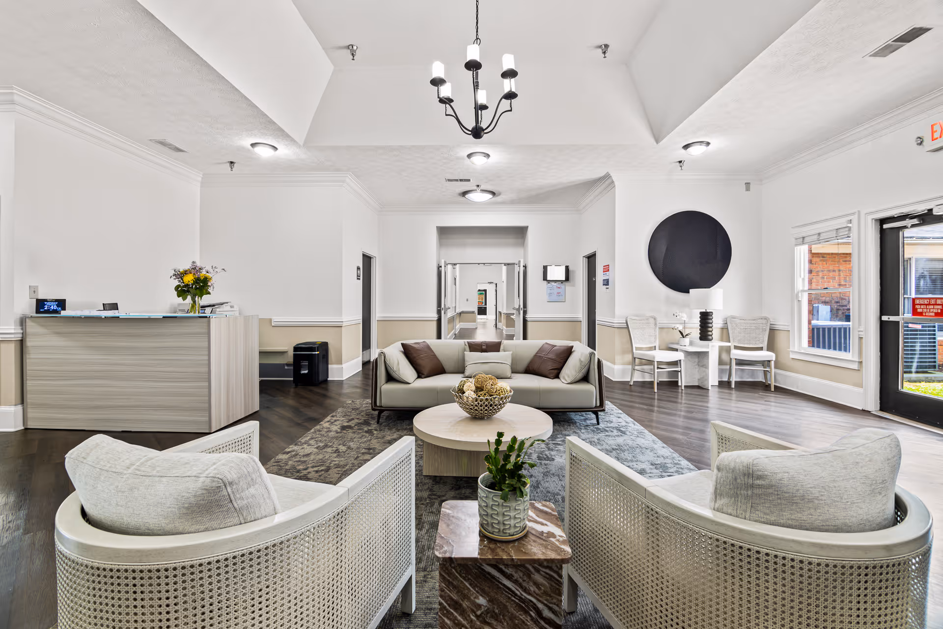A bright and modern assisted living facility lobby with a beige sofa and two cushioned chairs arranged around a round coffee table with decorative items. There is a reception desk with flowers on the left side, a small table with two chairs and a lamp on the right, and a hallway leading further into the building. The walls are white with beige wainscoting, and the floor is dark wood with area rugs. A black chandelier hangs from the ceiling.