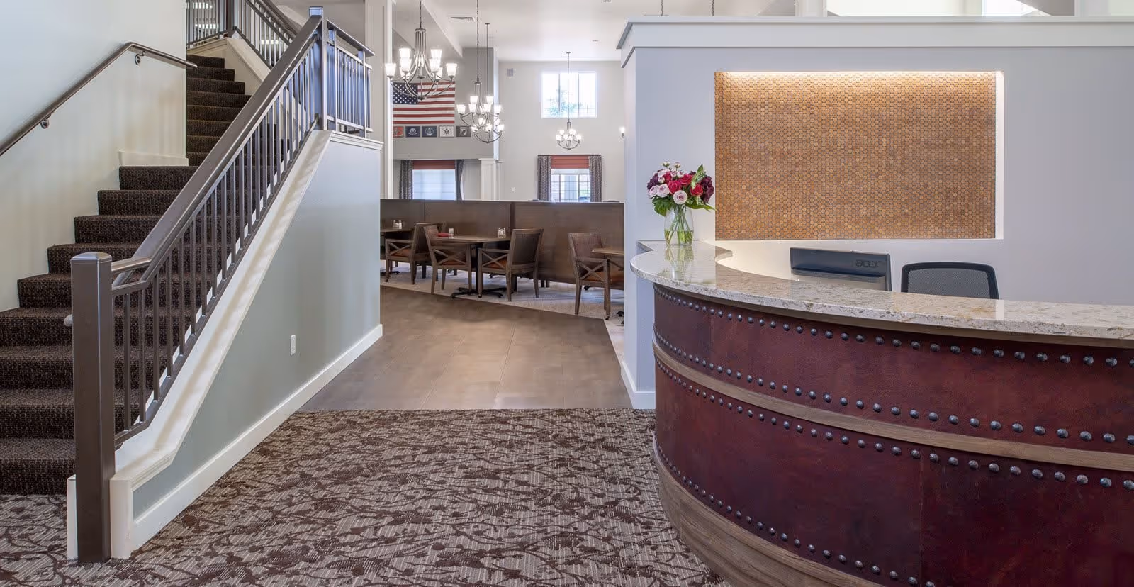 Interior view of a senior living facility showing a curved reception desk with a granite countertop and a vase of flowers. To the left, there is a carpeted staircase with a metal railing. In the background, there is a dining area with tables and chairs, chandeliers, and an American flag on the wall.
