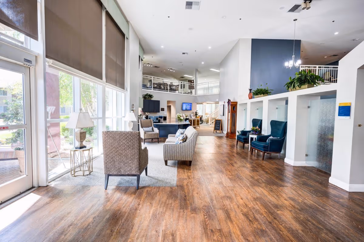 Bright and spacious senior living facility common area with large windows on the left letting in natural light. The room features wood flooring, several armchairs and sofas arranged for conversation, side tables with lamps, and plants placed on ledges. There is a second-floor balcony with railings visible in the background and a reception or service desk area further inside.