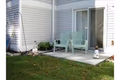 Small outdoor patio area with two light green plastic chairs on a concrete slab outside a sliding glass door of a white siding building. There is a small garden bed with plants and rocks adjacent to the patio, and a grassy lawn in the foreground.