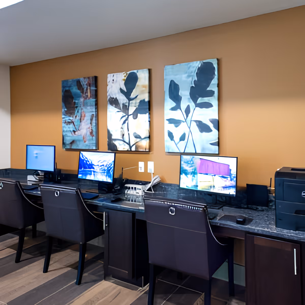 A workspace area with three computer stations on a dark granite countertop against a tan wall. Each station has a black chair and a computer monitor. Above the countertop, there are three abstract paintings featuring leaf designs. A printer is visible on the right side of the countertop.