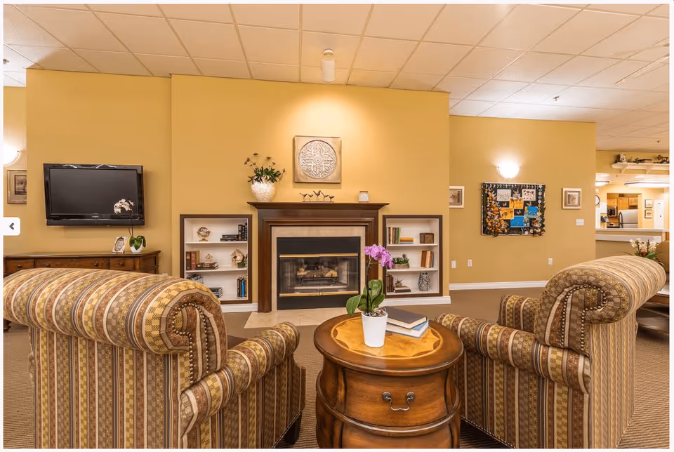 A cozy senior living facility common area with two patterned armchairs facing a wooden round table with a potted orchid and books. Behind the table is a fireplace with built-in shelves on either side containing books and decorative items. A flat-screen TV is mounted on the left wall above a wooden cabinet. The walls are painted yellow, and there is a bulletin board with photos and notes on the right wall. The ceiling has recessed lighting and a drop ceiling design.