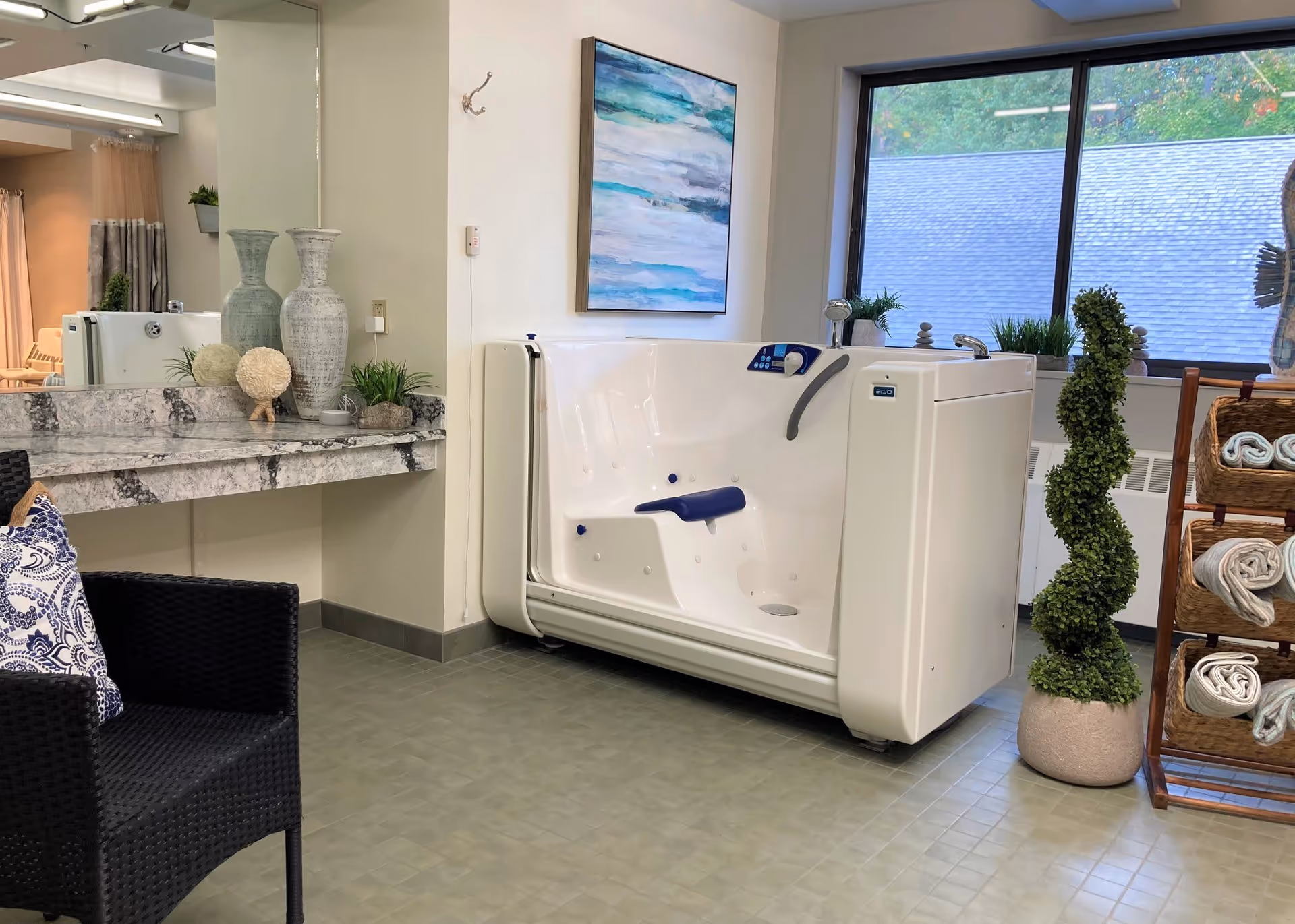 A clean and well-lit bathing area featuring a white walk-in bathtub with a blue seat and control panel. The room has a large window with a view of a roof and greenery outside. There is a decorative plant in a pot next to the tub, a wooden rack holding rolled towels, a black woven chair with a blue and white patterned cushion, and a marble countertop with decorative vases and plants. A large mirror and a colorful abstract painting adorn the walls.