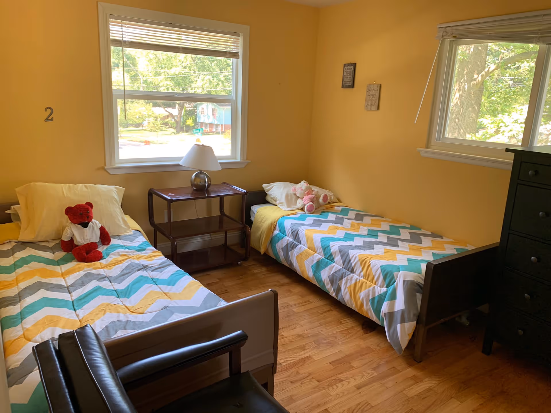 A bright bedroom with yellow walls featuring two single beds with colorful zigzag patterned bedspreads. One bed has a red teddy bear and the other has a pink teddy bear. There is a wooden nightstand with a lamp between the beds, two windows letting in natural light, and a black dresser on the right side of the room.