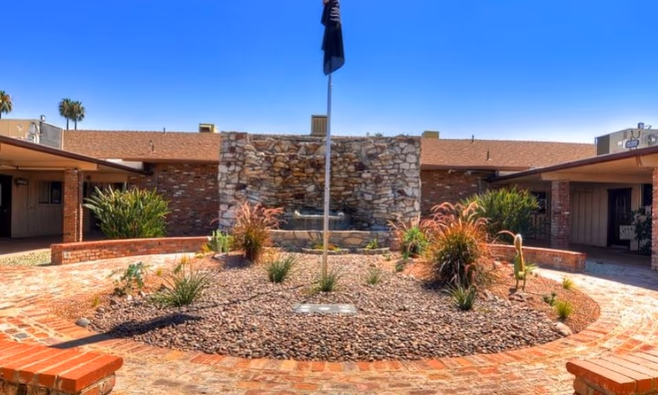 Circular brick courtyard with a central flagpole and desert landscaping in front of a stone fountain wall and single-story brick building under a clear blue sky.