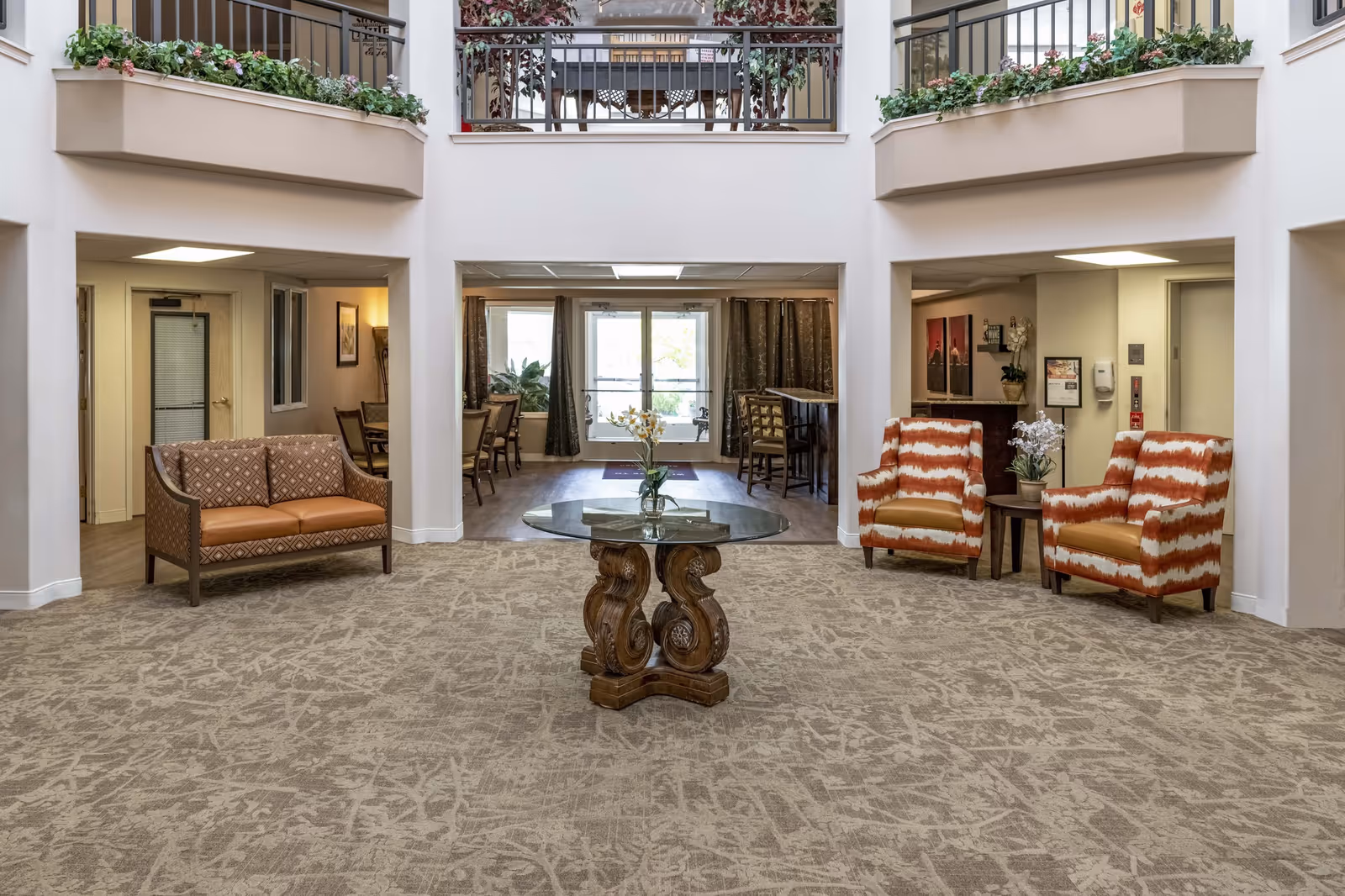 Spacious and well-lit interior common area of a senior living facility with patterned carpet, a round glass-top table with a floral arrangement in the center, two orange and white patterned armchairs with a small side table and flowers between them on the right, and a brown patterned loveseat on the left. The area opens into a dining space with tables and chairs, large windows, and a door leading outside. The upper level features railings with flower boxes filled with greenery and flowers.