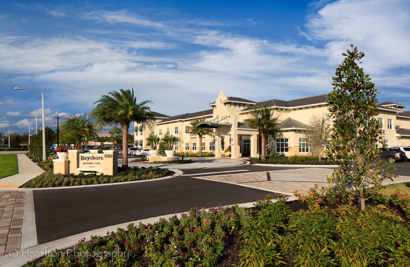 Exterior view of Bayshore Memory Care facility showing a large beige building with multiple windows, a covered entrance, palm trees, landscaped flower beds, and a paved driveway under a partly cloudy blue sky.