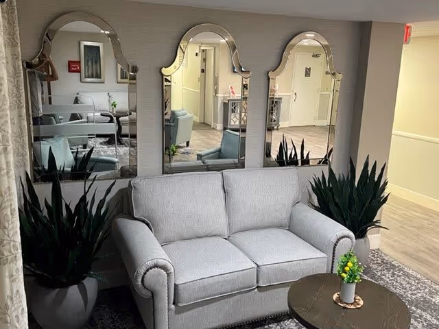 A cozy seating area in a senior living facility featuring a light gray loveseat with rolled arms, flanked by two large potted plants. Behind the loveseat are three decorative mirrors with ornate tops mounted on the wall. In front of the loveseat is a round wooden coffee table with a small potted plant on it. The background shows a hallway with light-colored walls and flooring.