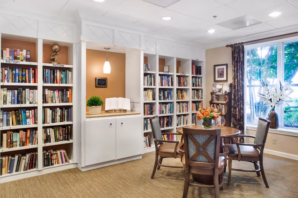 A cozy library room with white bookshelves filled with books labeled 'Fiction'. There is a round wooden table with four cushioned chairs around it. A vase with flowers is placed on the table. A large window with floral curtains lets in natural light, and a decorative plant and an open book are displayed on a built-in cabinet under a hanging light fixture.