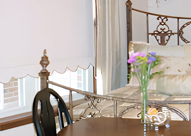 Wooden table with a vase of purple flowers in front of an ornate metal bed and window with blinds.