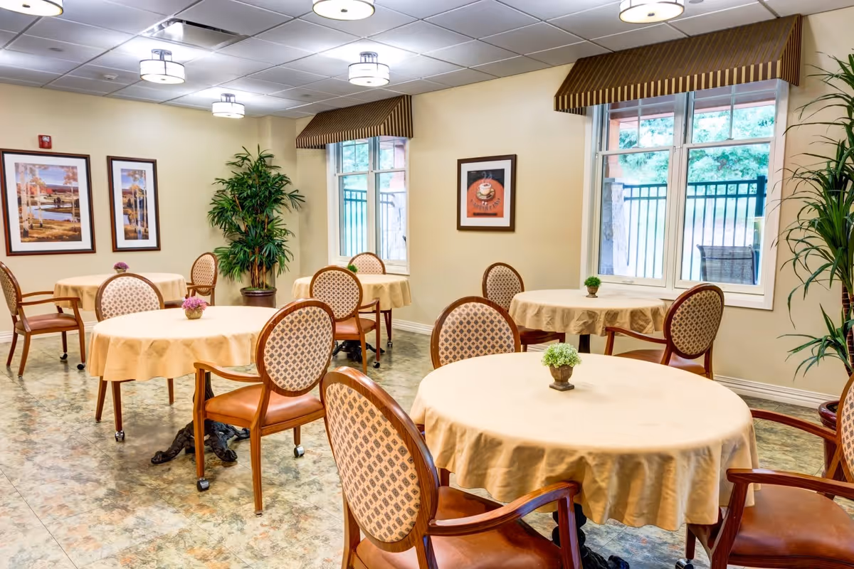 A bright dining room with round tables covered in beige tablecloths, each table adorned with a small potted plant. The room features patterned chairs with wooden frames, large windows with striped valances, framed artwork on the walls, and potted plants in the corners.