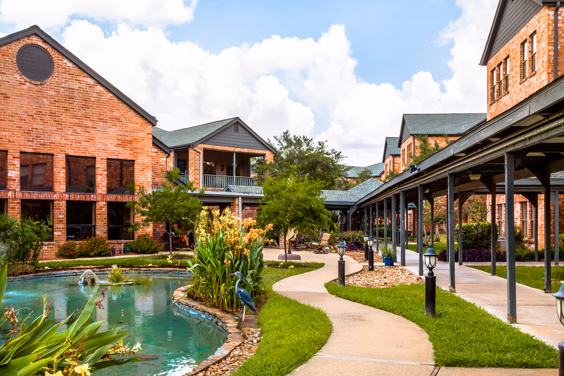 Outdoor courtyard area at Tarrytowne Estates featuring a curved walkway, a pond with a small fountain, lush greenery, and brick buildings with covered walkways under a partly cloudy sky.