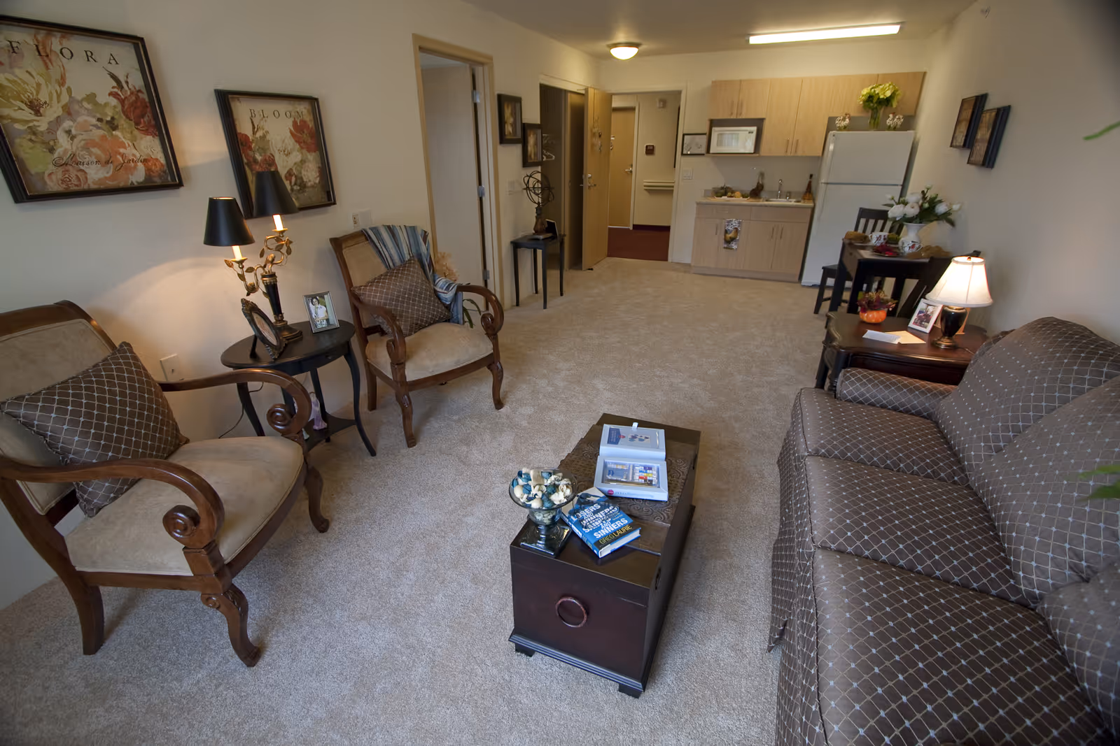 A cozy senior living room with beige carpet, a patterned brown sofa, two wooden armchairs with cushions, a small black coffee table with books and decorative items, a side table with a lamp and framed photos, and a kitchenette with light wood cabinets, a microwave, and a refrigerator in the background.
