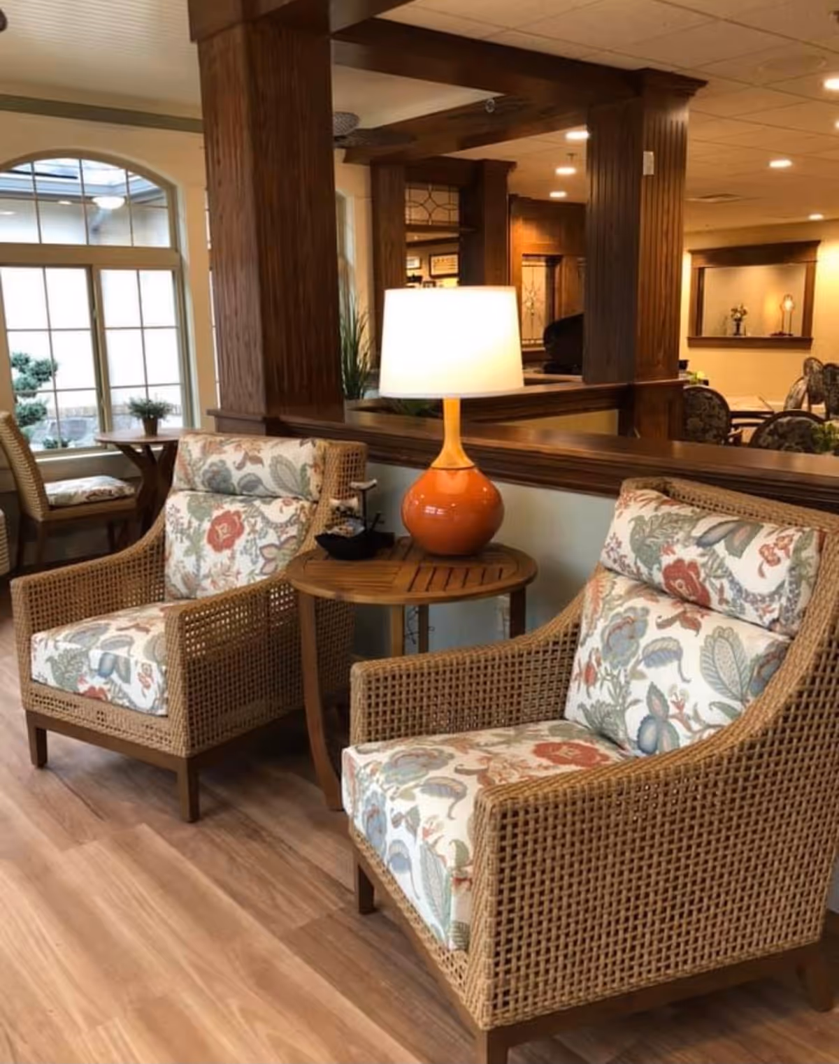 Two wicker armchairs with floral cushions flanking a small round table topped by an orange lamp in a warm communal lounge area.