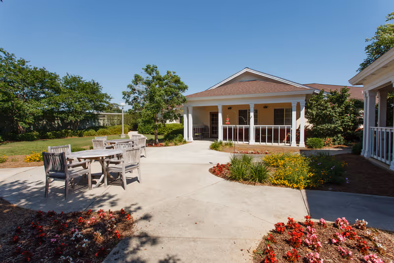 Outdoor patio area at The Brennity at Daphne Assisted Living & Memory Care featuring wooden tables and chairs on a concrete surface surrounded by flower beds and greenery, with a building porch in the background under a clear blue sky.