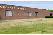 Exterior view of a single-story brick building with the sign 'ORCHARD MANOR' on the wall. There are three windows visible and a well-maintained grassy area in front of the building under a clear sky.