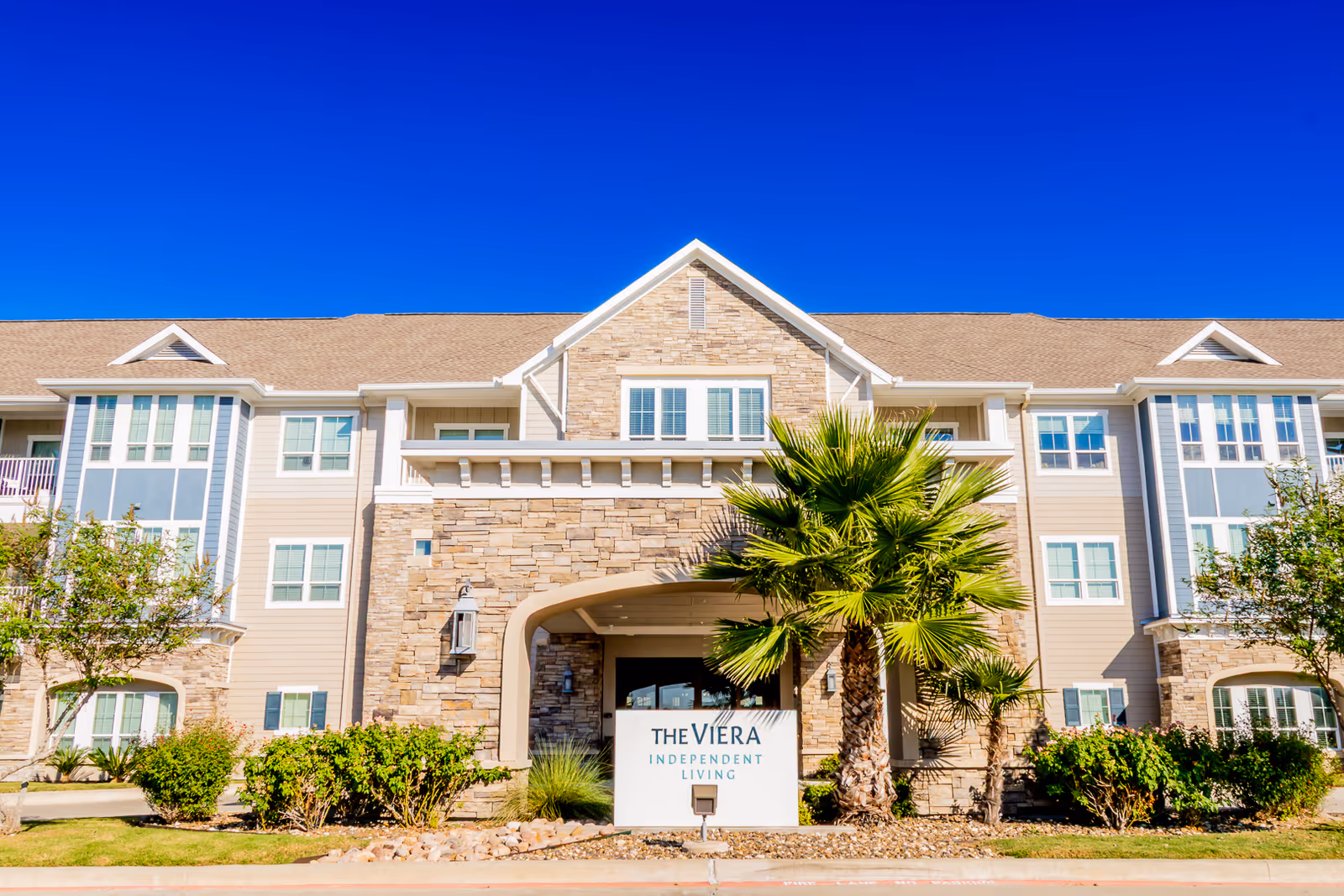 Exterior front view of The Viera Senior Living facility showing a multi-story building with beige and stone facade, large windows, and a sign reading 'The Viera Independent Living' surrounded by palm trees and shrubs under a clear blue sky.