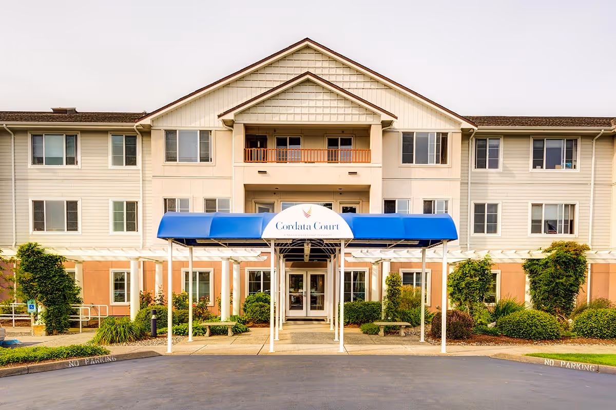 Front entrance of a three-story senior living building with a blue awning that reads "Cordata Court" and landscaped grounds.