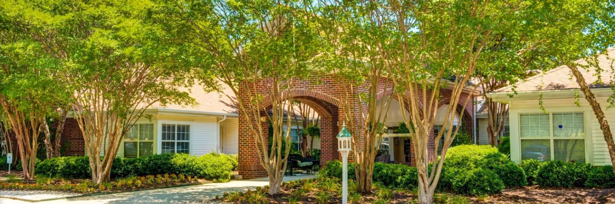 Exterior view of a senior living facility with a brick archway entrance surrounded by green trees and bushes. The building has white siding and multiple windows with white shutters. A paved walkway leads to the entrance, and there is a white post with a small lantern in the landscaped area.