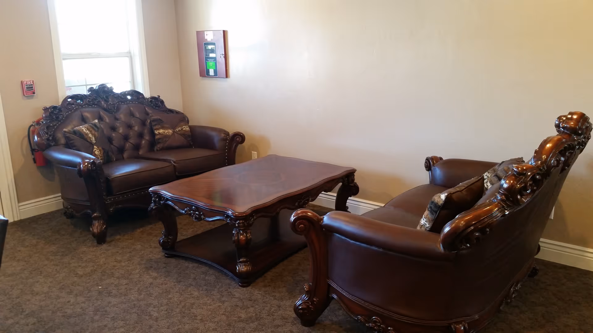 A sitting area with two ornately carved dark brown leather sofas facing each other and a matching wooden coffee table in between. The room has beige walls, a window with white trim, and a fire alarm and fire extinguisher mounted on the wall near the window.