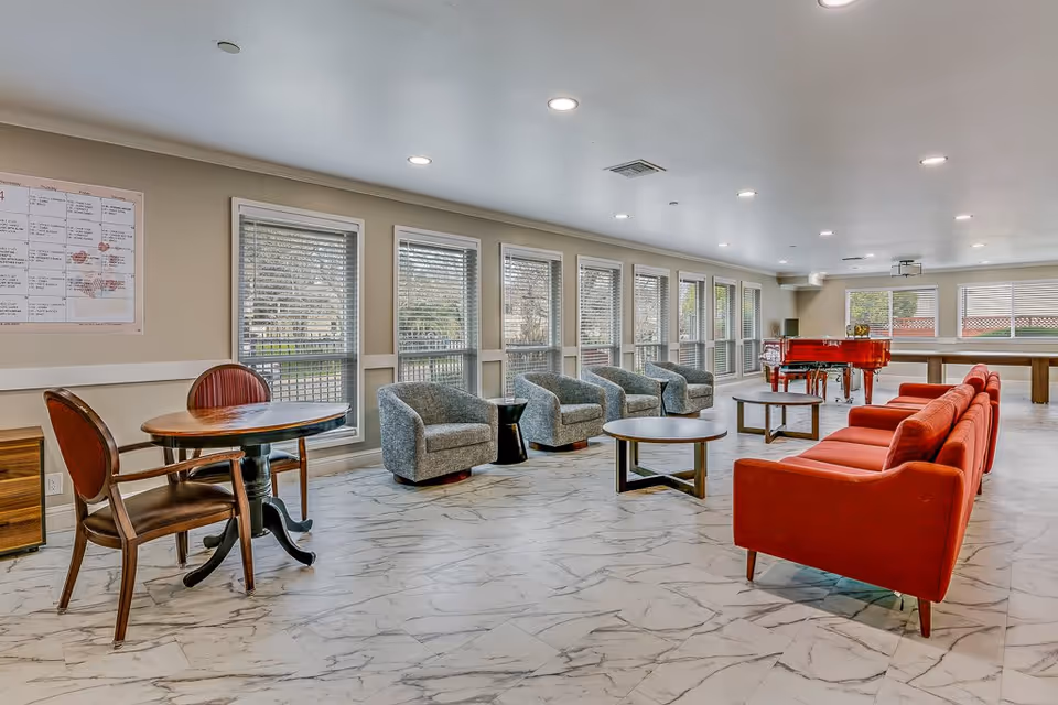 A spacious and well-lit common area in an assisted living facility featuring a row of large windows with blinds, several gray armchairs, a red sofa, round wooden tables, a small table with two chairs, and a red piano in the background.