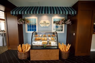 A bakery display area inside a facility with a glass case filled with various baked goods. Two baskets filled with baguettes are placed on either side of the display. The backdrop features a sign that reads 'The Bakery' with two windows and hanging flower baskets under a striped awning.