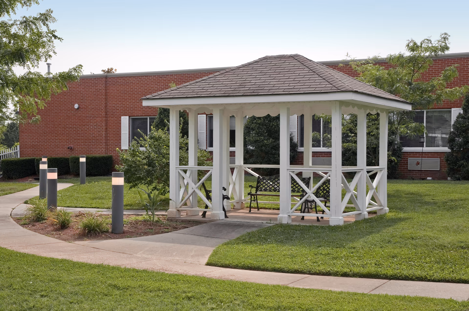 A white wooden gazebo with a shingled roof situated on a grassy lawn with a concrete pathway leading to it. The gazebo contains two black metal benches and is surrounded by green trees and shrubs. In the background, there is a red brick building with windows and white shutters.
