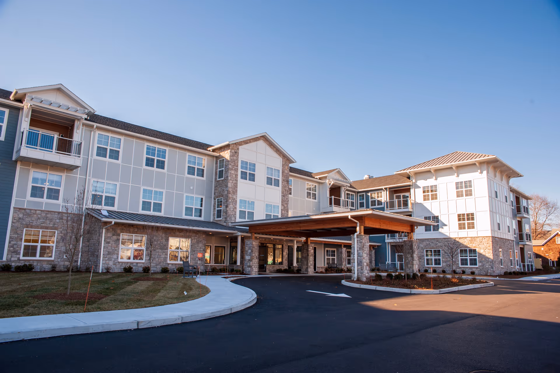 Exterior view of a multi-story senior living facility building with stone and light-colored siding, large windows, and a covered entrance driveway under a clear blue sky.
