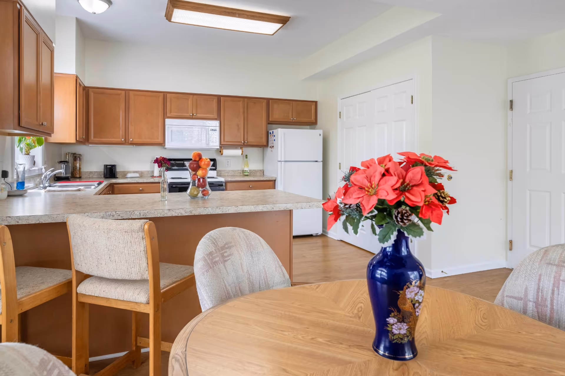Bright kitchen and dining area with wooden cabinets, a breakfast bar, and a vase of red flowers on the table.