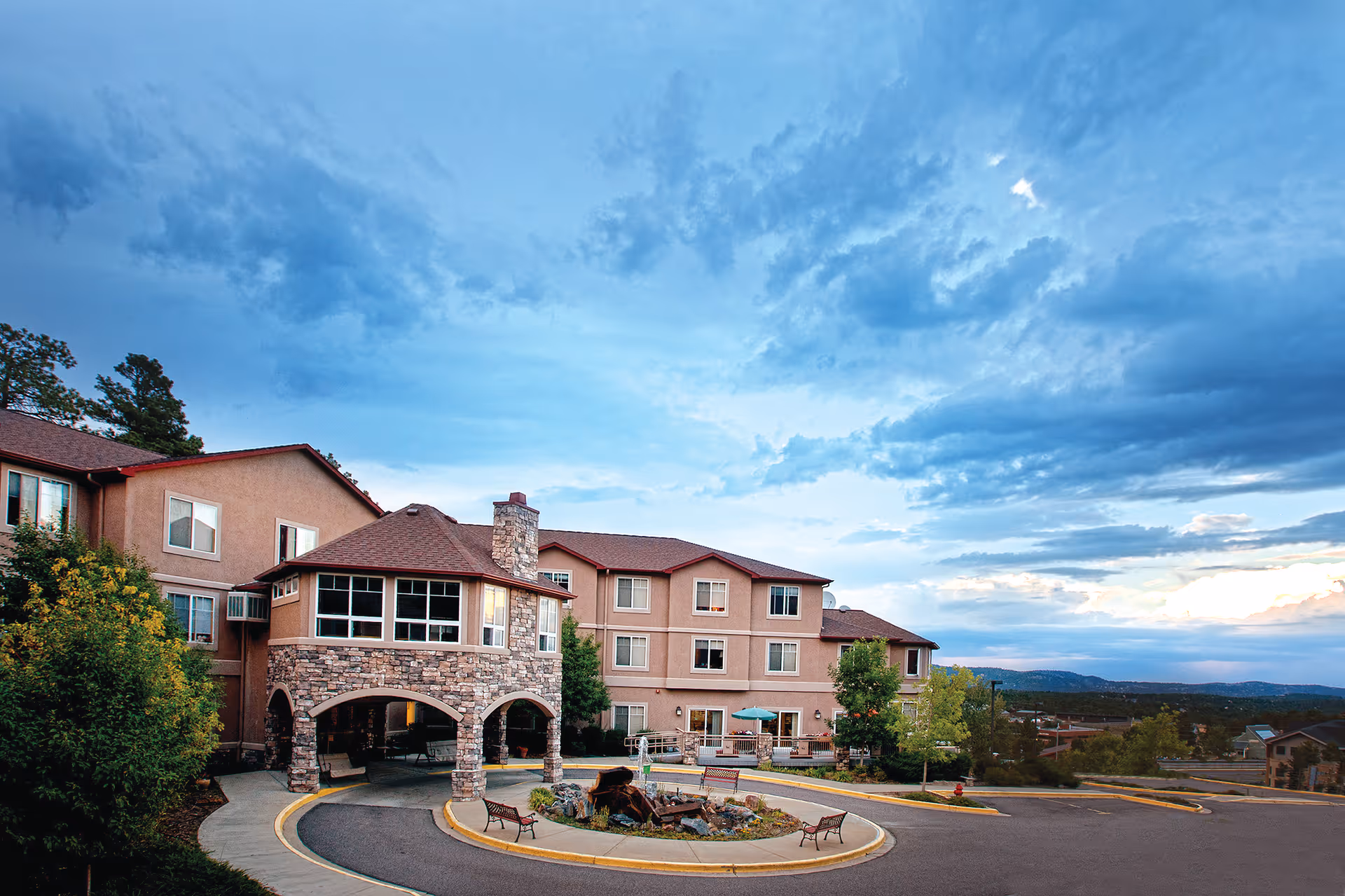 Front exterior of a multi-story assisted living building with a stone arched entry and circular driveway under a cloudy sky.