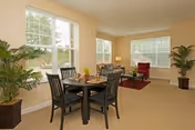 Open, sunlit living and dining area with a round dining table and chairs in the foreground and a seating area with an armchair and coffee table by large windows.