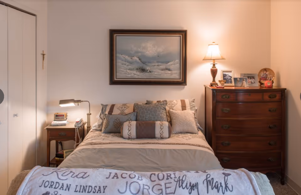 Tidy bedroom with a made bed topped with multiple decorative pillows, a nightstand and lamp on the left, and a wooden dresser with a lamp and framed photos on the right.