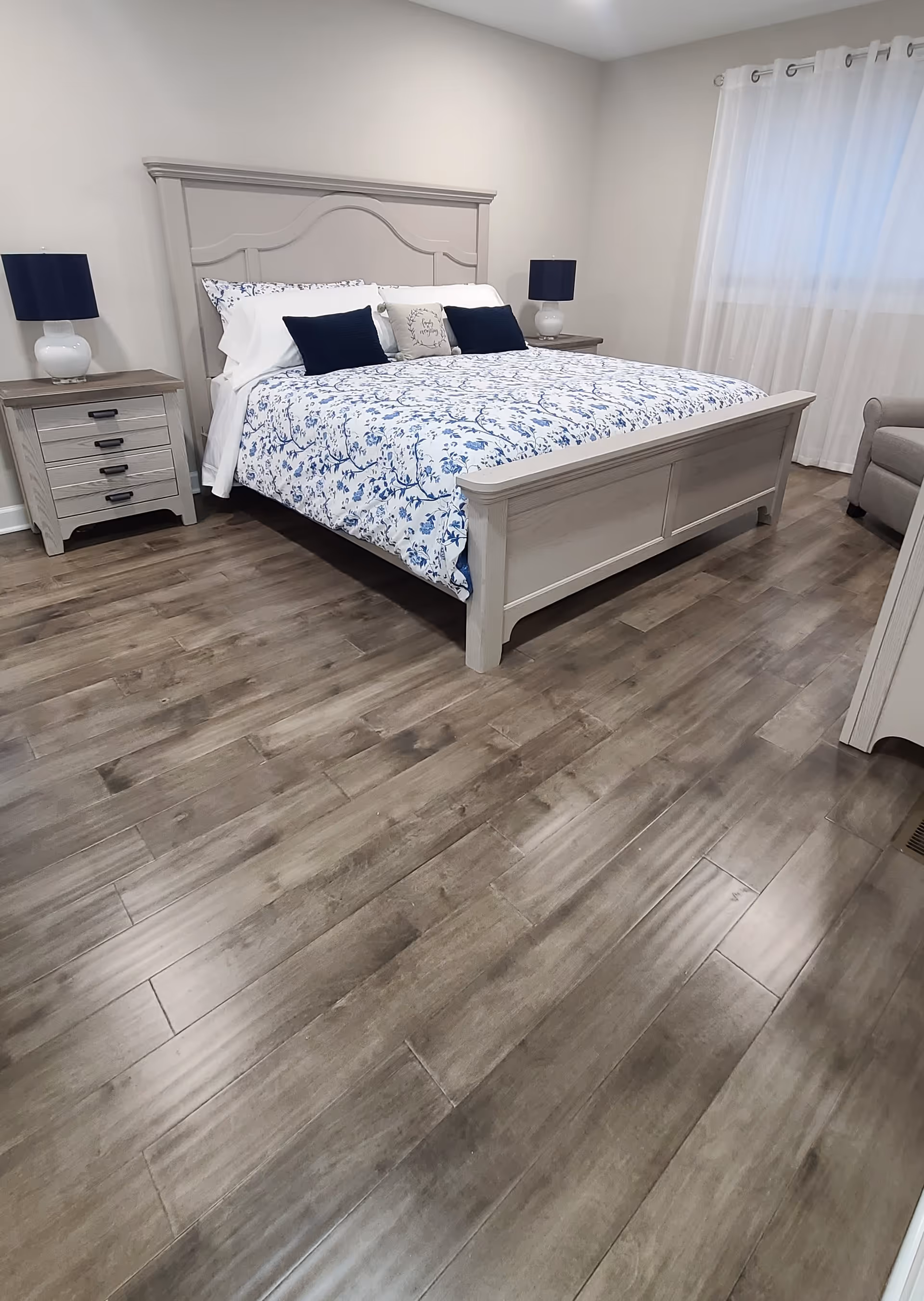 A bedroom with a large bed featuring a light gray wooden frame and a floral blue and white bedspread. There are two matching nightstands on either side of the bed, each with a white lamp with a dark blue lampshade. The room has light-colored walls, a window with white curtains, and wood-look flooring.