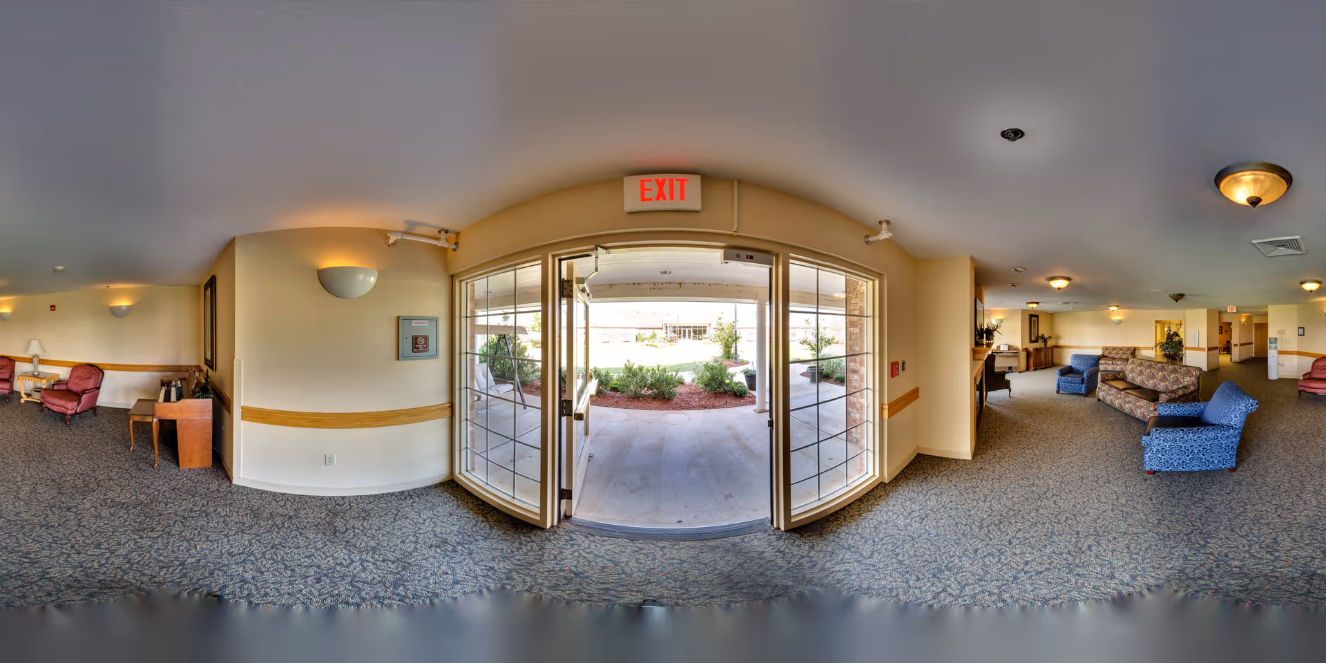 Panoramic view of a nursing center's interior common area with carpeted floor, multiple seating arrangements including armchairs and sofas, wall-mounted lights, and an exit door leading outside to a garden area.